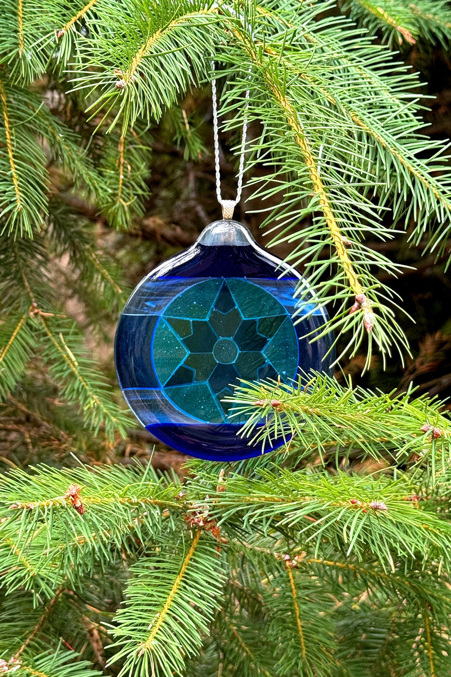 Blue 6-Point Star Fused Glass Ornament by AWinburnART hanging on a pine tree branch.