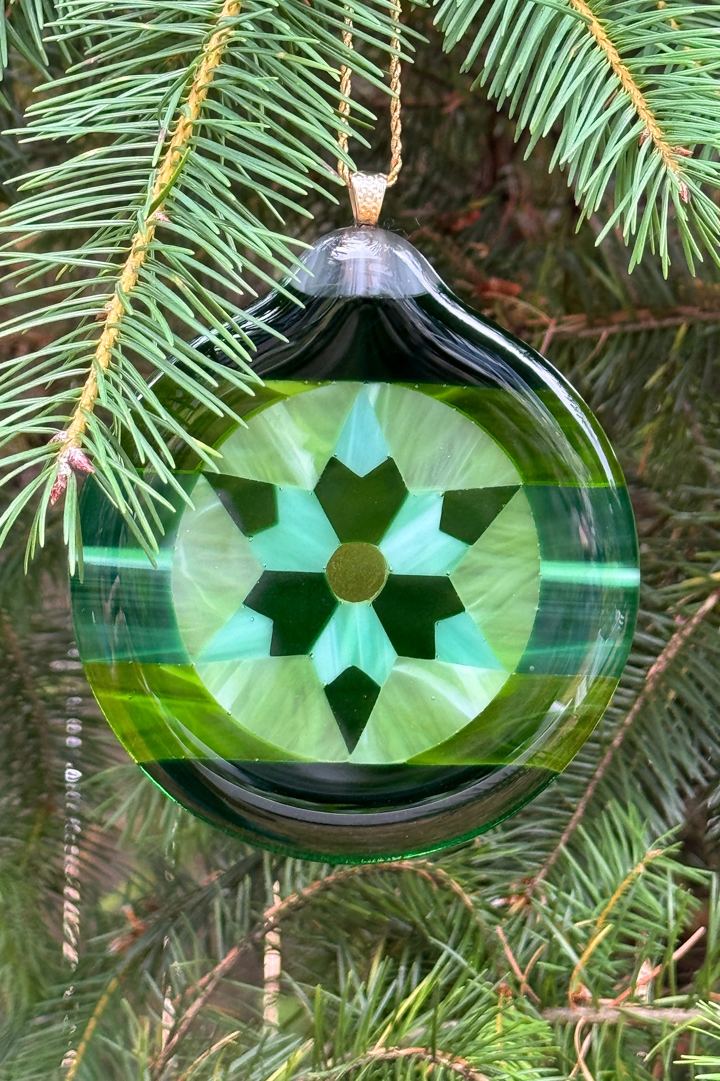 A round handcrafted green glass Christmas ornament with a geometric star pattern, hanging from a gold loop on a pine tree branch by AWinburn.
