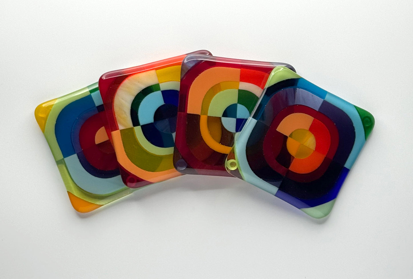 Four Squarckles fused glass coasters fanned in a stack showing multicolor hard-edge concentric circle patterns in stained art glass, handcrafted and signed by AWinburnART