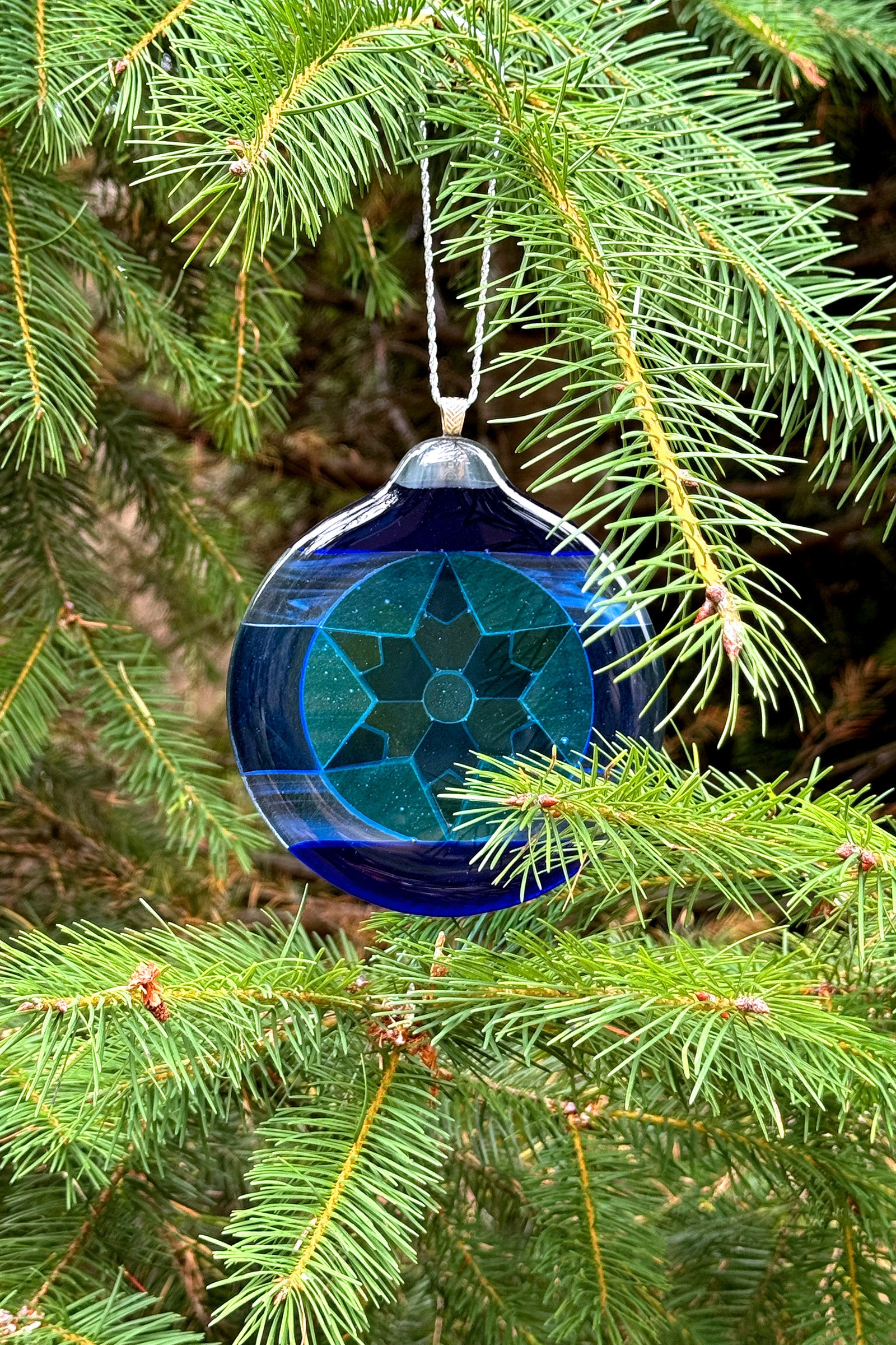 Blue 6-Point Star Fused Glass Ornament by AWinburnART hanging on a pine tree branch.