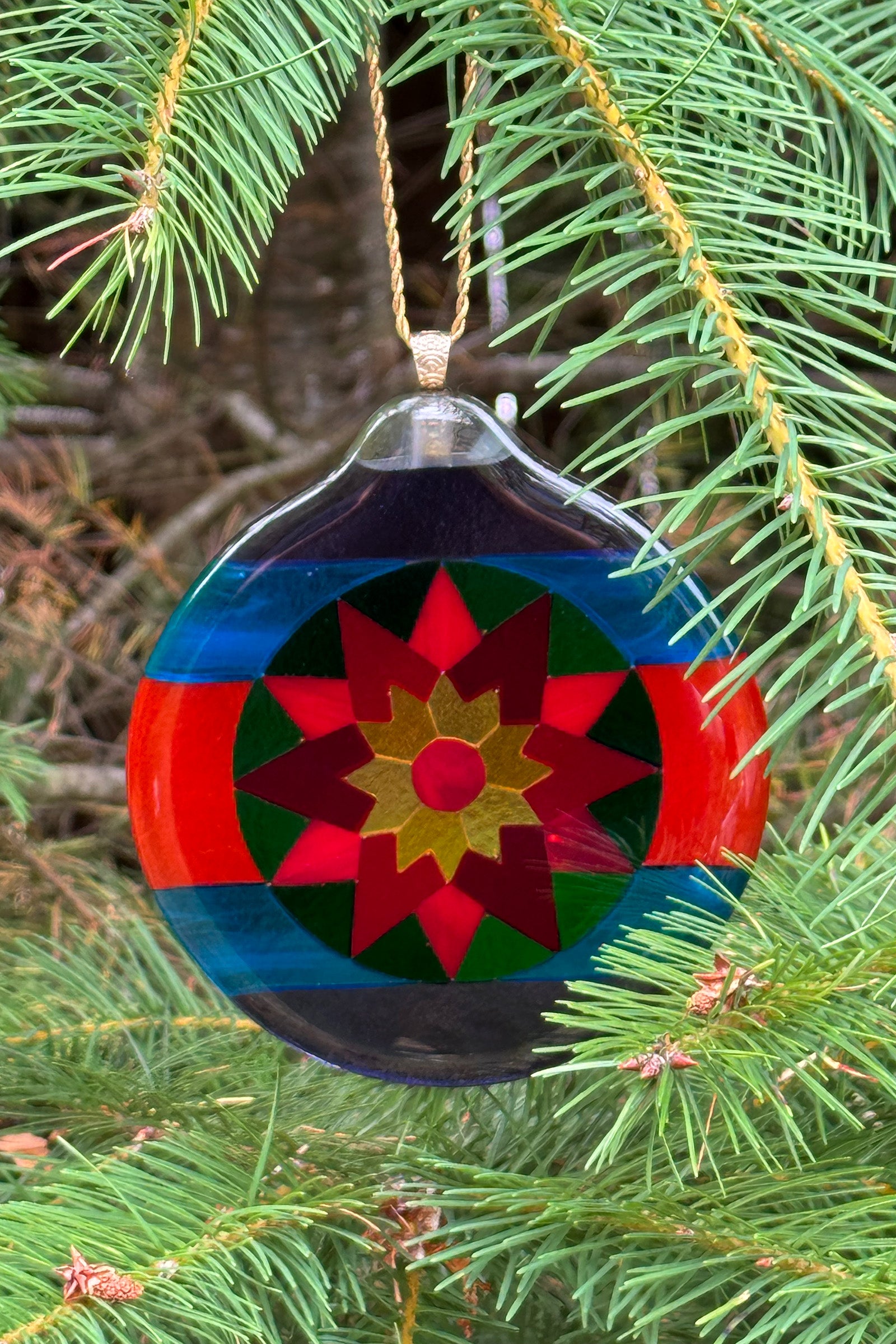 Fair Isle Poinsettia fused glass ornament hanging on a pine Christmas tree, displaying rich red and green poinsettia pattern with yellow center.