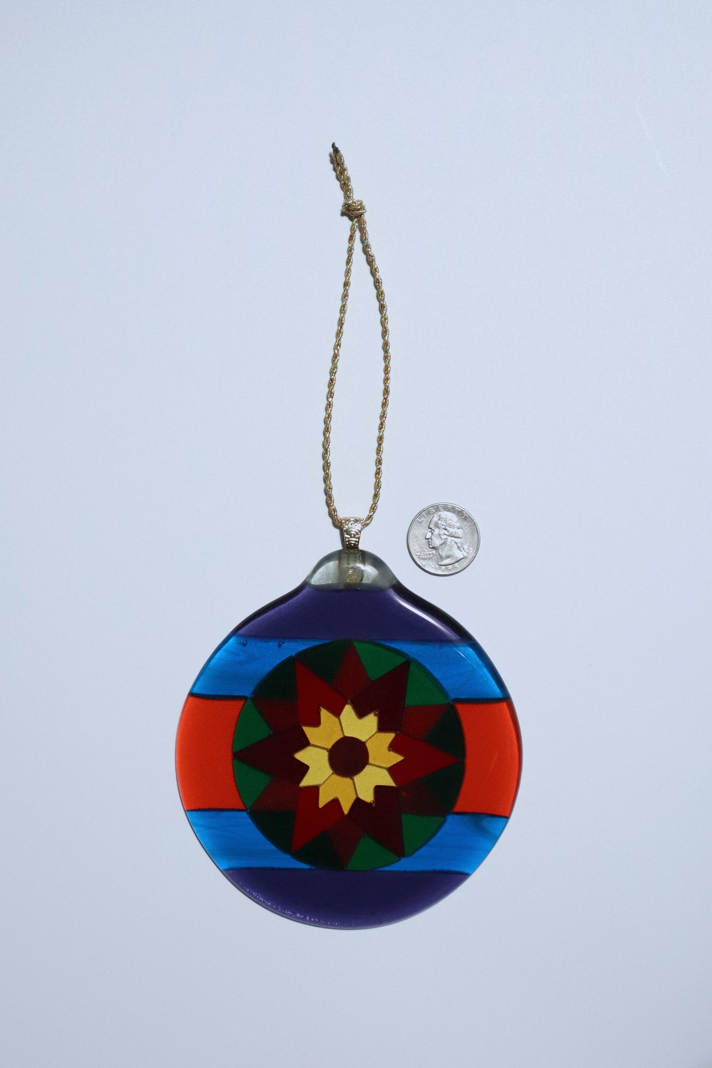Round Fair Isle Poinsettia fused glass ornament on a white background, showing bold geometric design in purple, blue red green, orang and yellow art glass with a quarter coin for scale.
