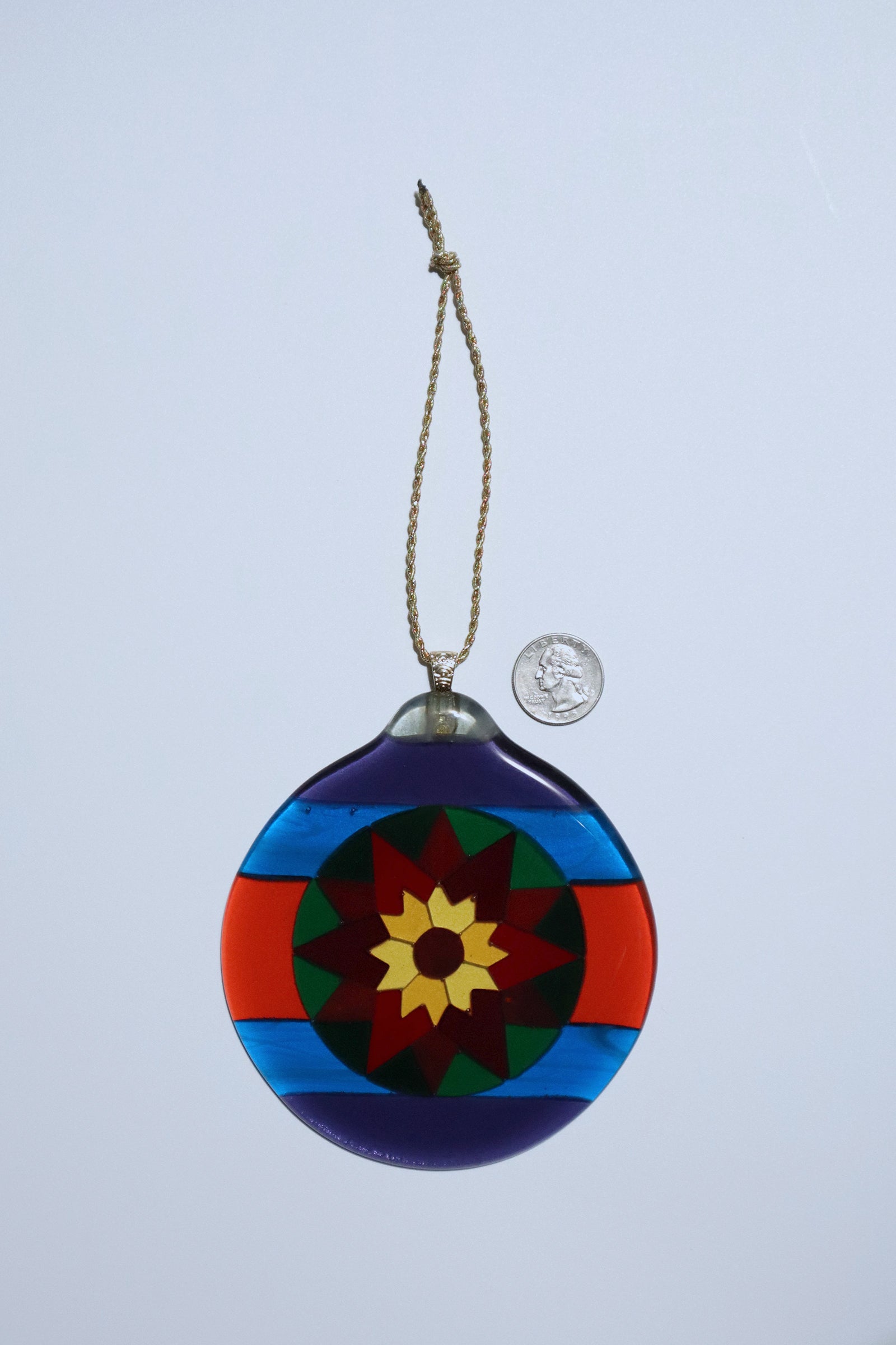 Round Fair Isle Poinsettia fused glass ornament on a white background, showing bold geometric design in purple, blue red green, orang and yellow art glass with a quarter coin for scale.