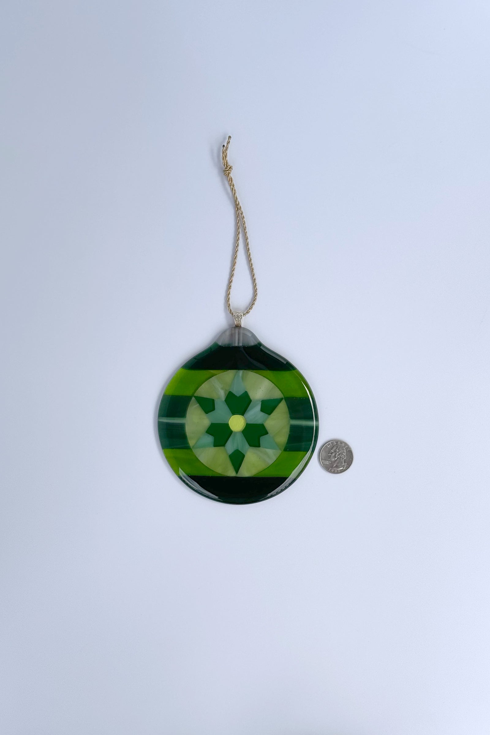A round fused glass hanging ornament with a green geometric star design, suspended by a gold-colored string next to a quarter coin for scale.