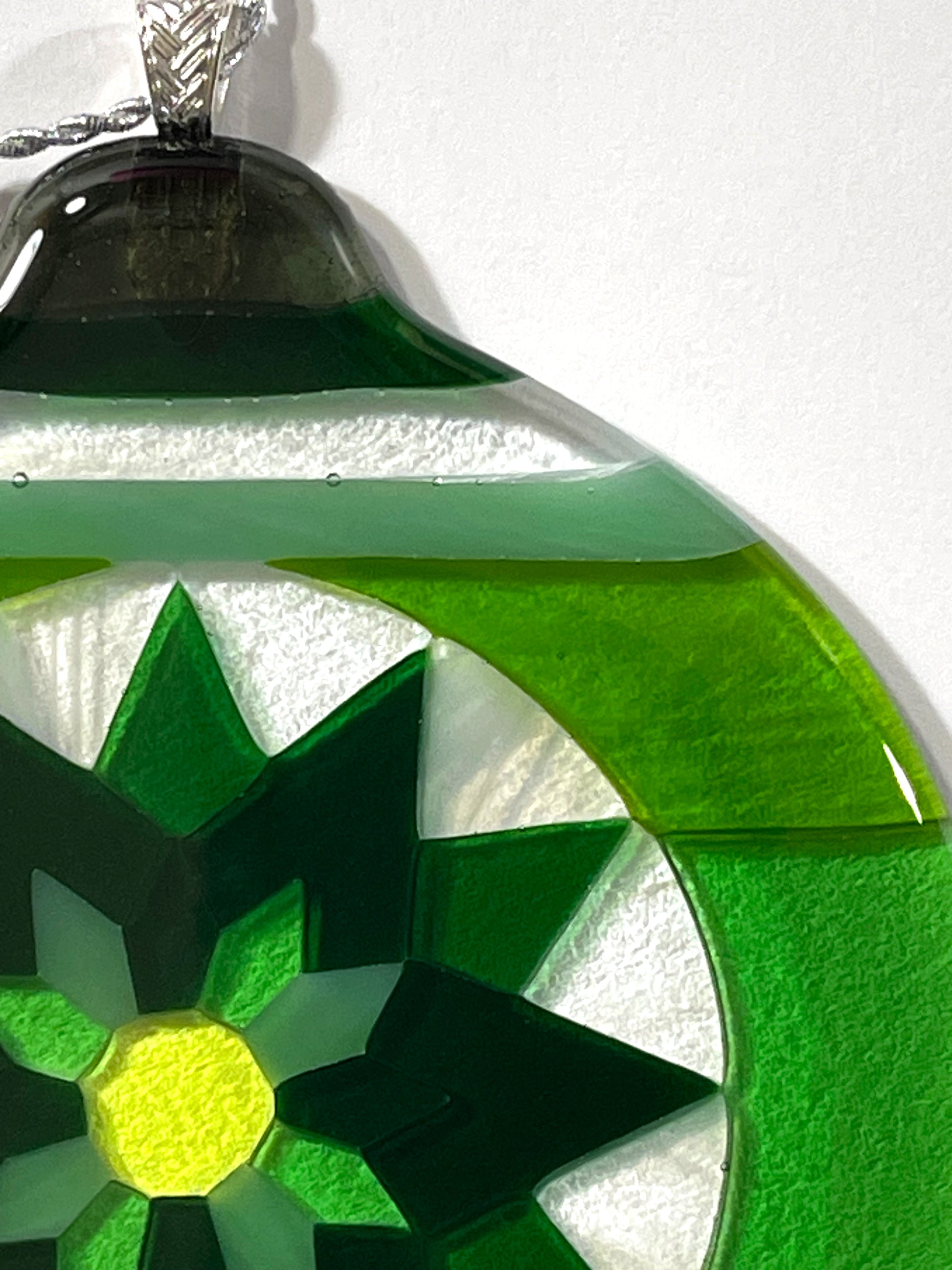 Close up detail of hand-cut art glass pieces in the Green Fair Isle Poinsettia fused glass ornament, showing dark green , bright green, and white wispy glass segments with a yellow-green center.
