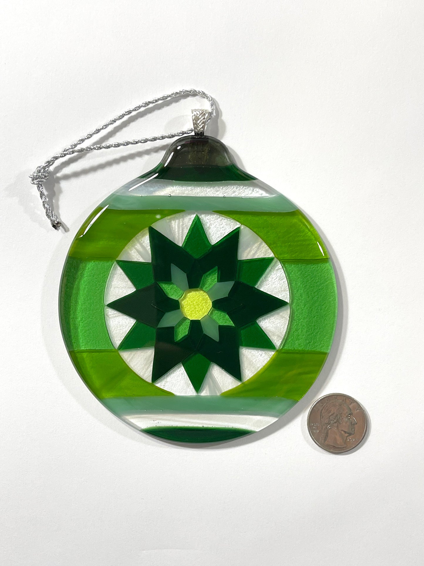 Green Fair Isle Poinsettia fused glass ornament shown with a quarter coin for scale, displaying geometric green star pattern with a yellow-green center.