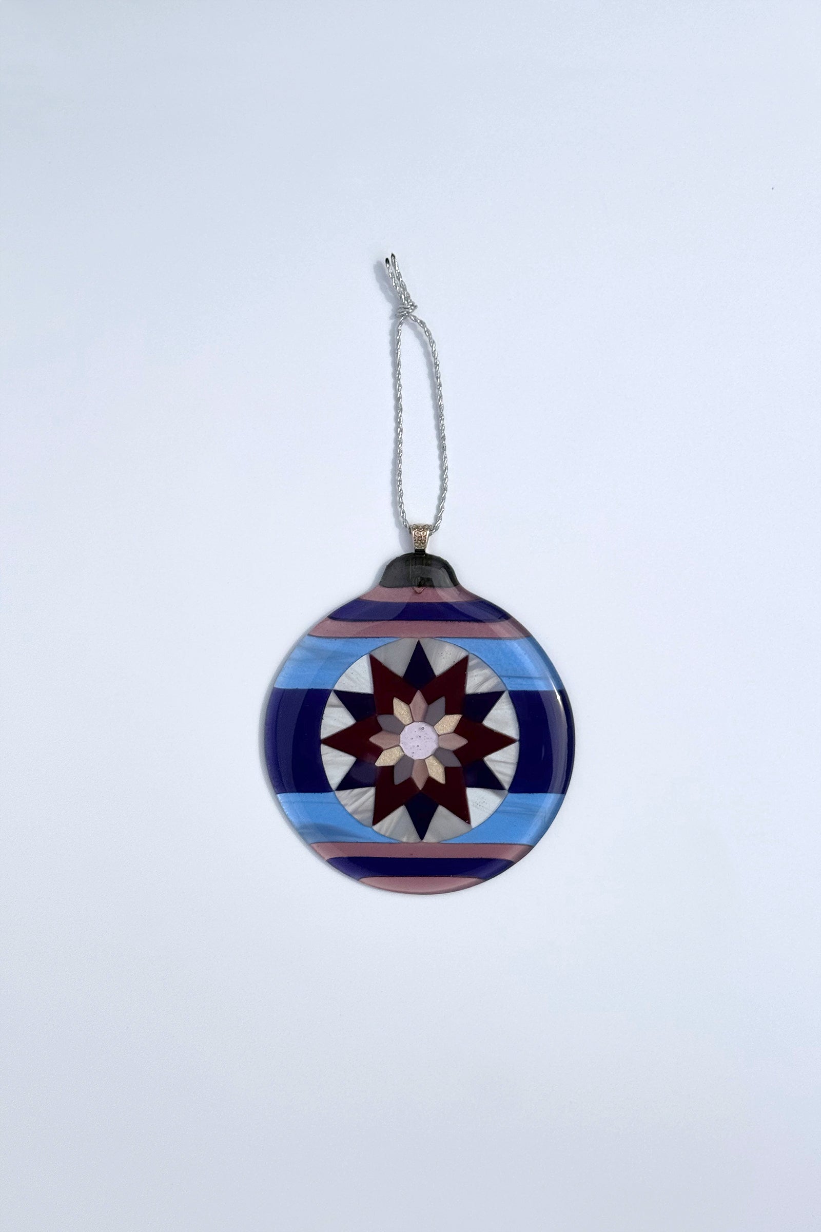 Fair Isle Poinsettia fused glass ornament in varying shades of purple, and blue with a sterling silver-plated bail and braided ribbon, against a white background.