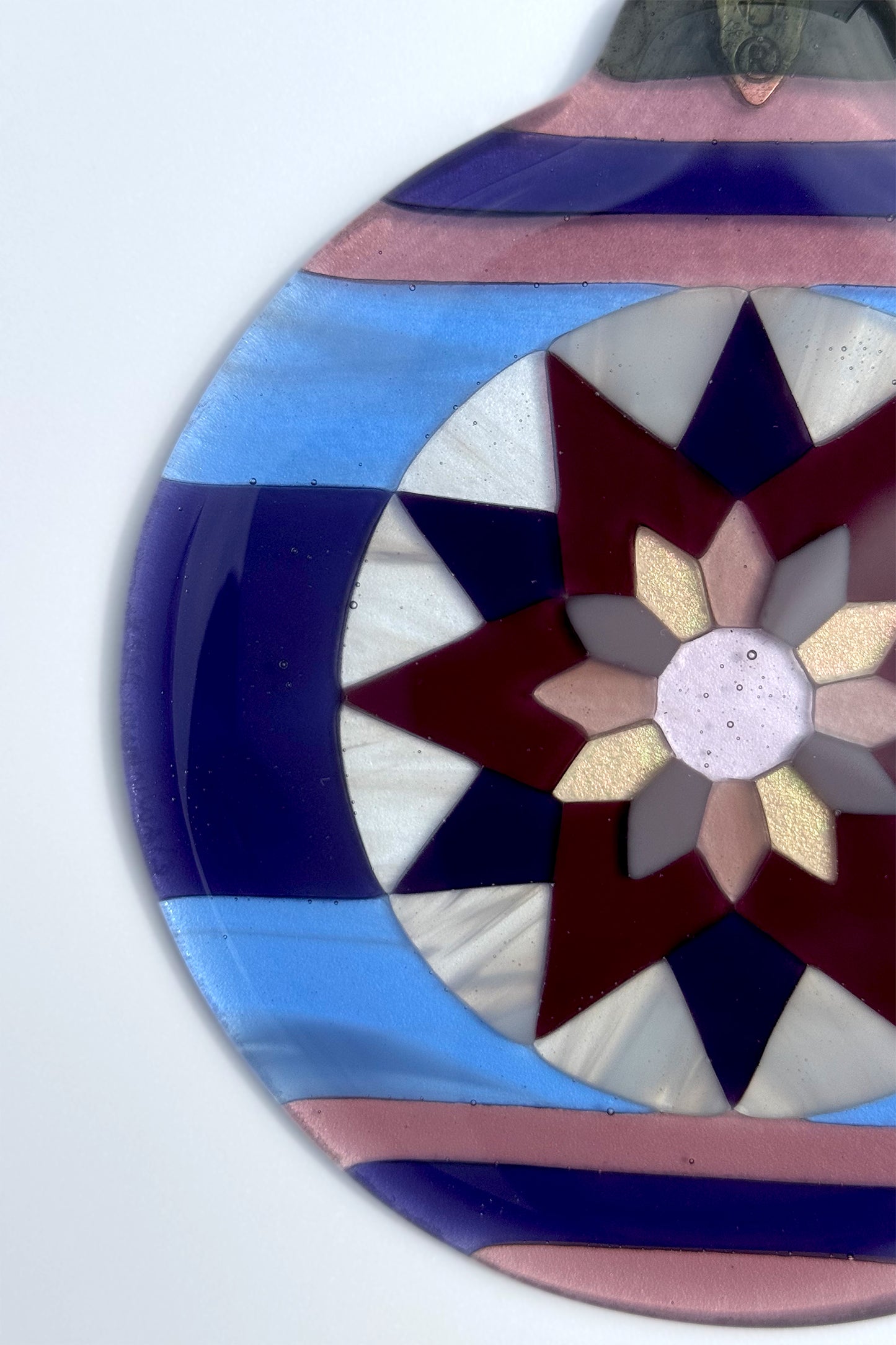 Close-up detail of hand cut glass pieces in varying shades of purple, blue, wispy white, and iridescent pink forming a Fair Isle poinsettia star pattern.