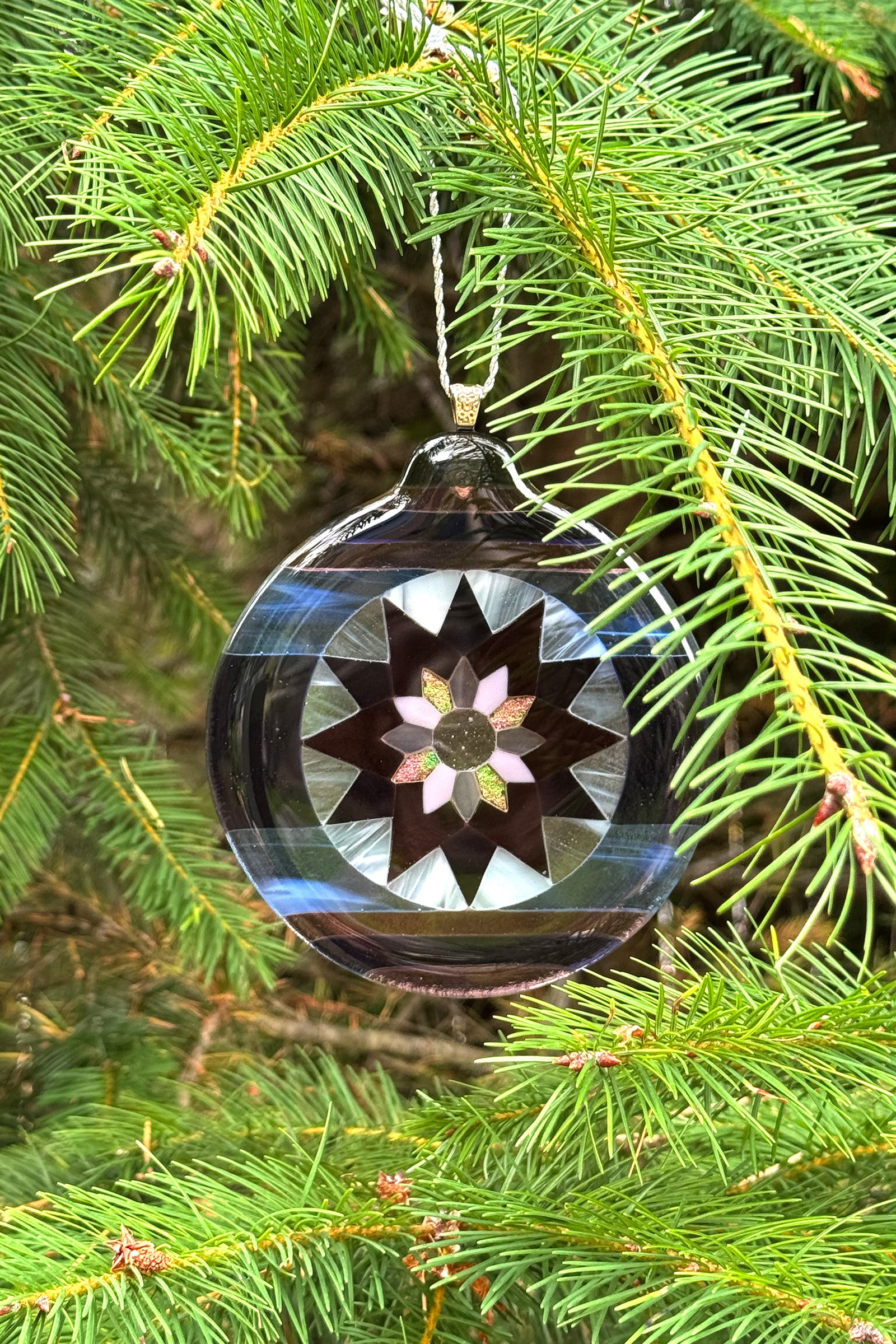 Handcrafted fused glass Fair Isle Poinsettia ornament hanging on a pine tree, showing translucent purple and blue glass while highlighting Iridescent pink in natural light.
