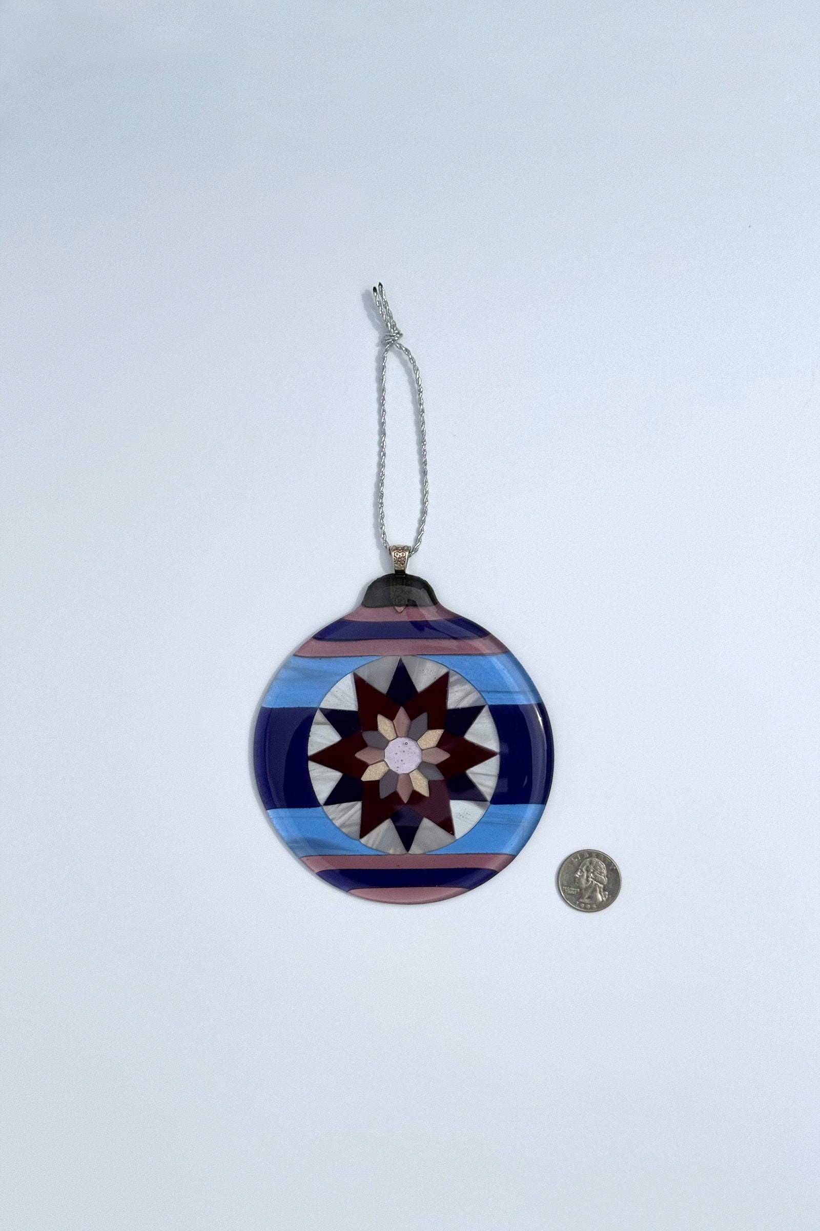 Fair Isle Poinsettia fused glass ornament in varying shades of purple and blue shown next to a quarter to indicate size, approximately 4½ inches wide.