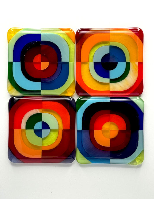Set of four Squarckles handcrafted fused glass coasters laid flat, each featuring bold concentric circle geometry in multicolor art glass — red, blue, orange, green, and yellow — by AWinburnART