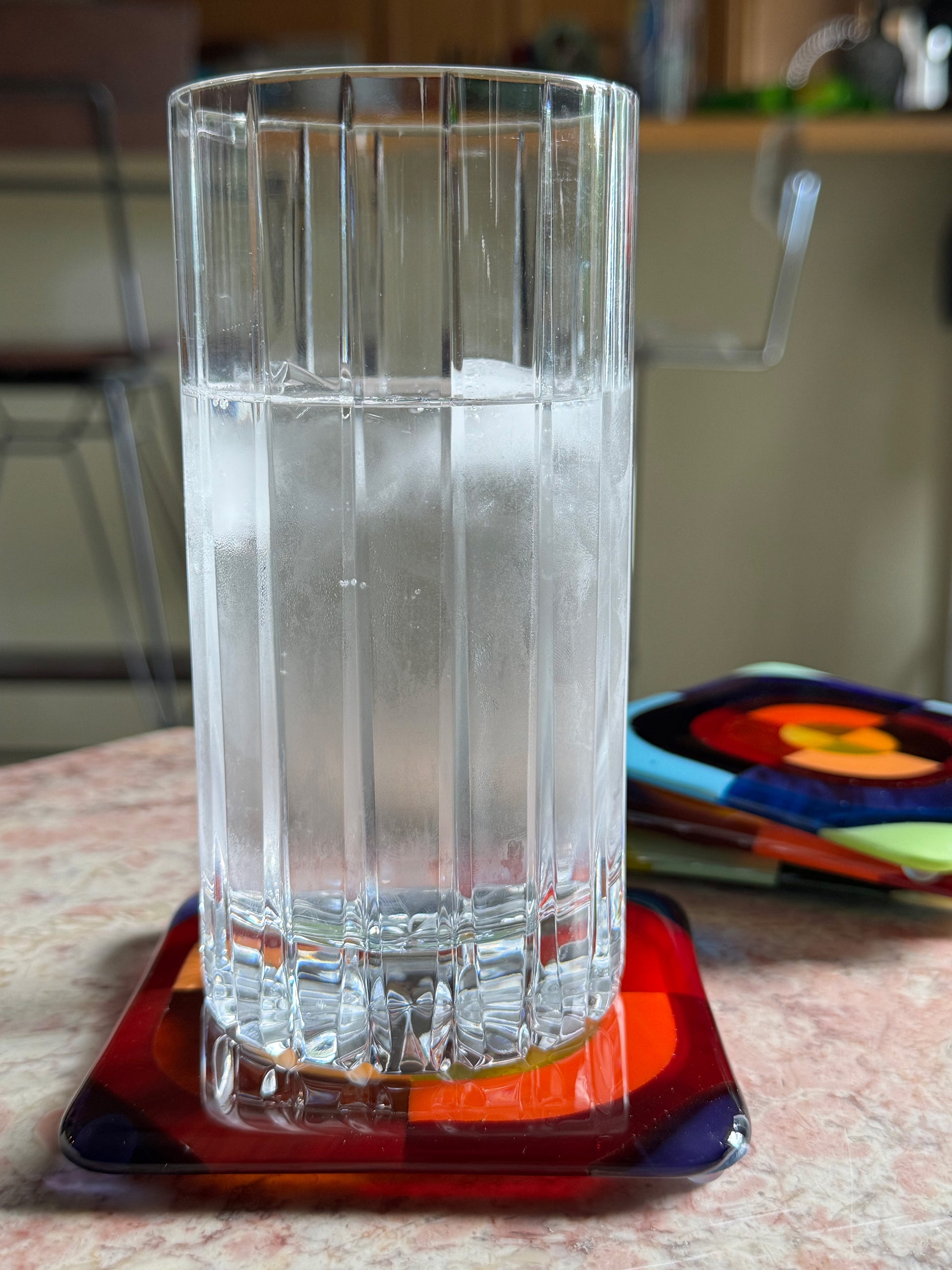Tall glass of water resting on a Squarckles fused glass coaster in deep red and orange concentric design, with remaining coasters stacked in background, AWinburnART